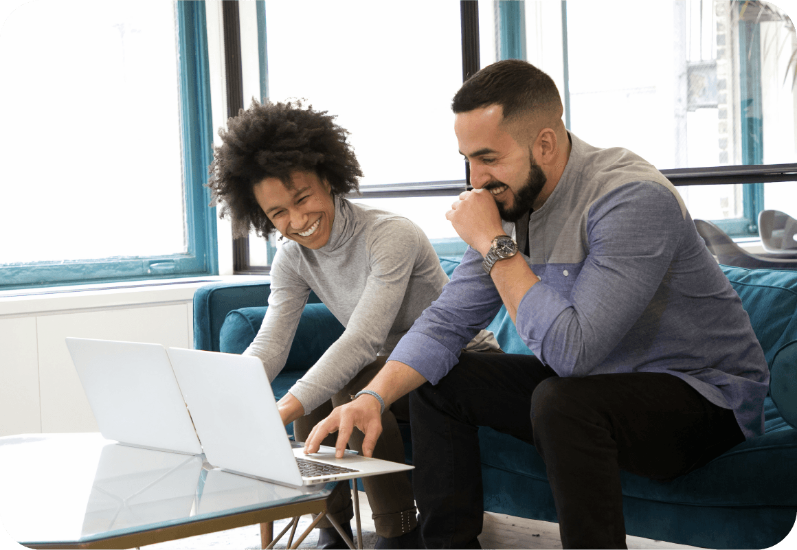 Zwei Personen sitzen auf einem blauen Sofa, lächeln und arbeiten gemeinsam an Laptops an einem gläsernen Kaffeetisch in einem hellen Büro.