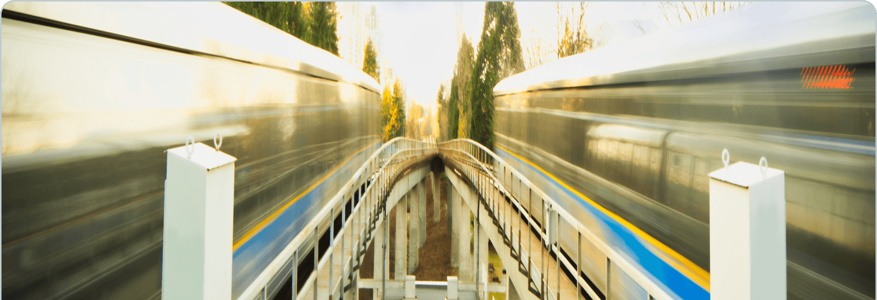 Due treni sfocati che sfrecciano su una piattaforma stretta con ringhiere bianche e strisce giallo-blu, alberi visibili sullo sfondo.