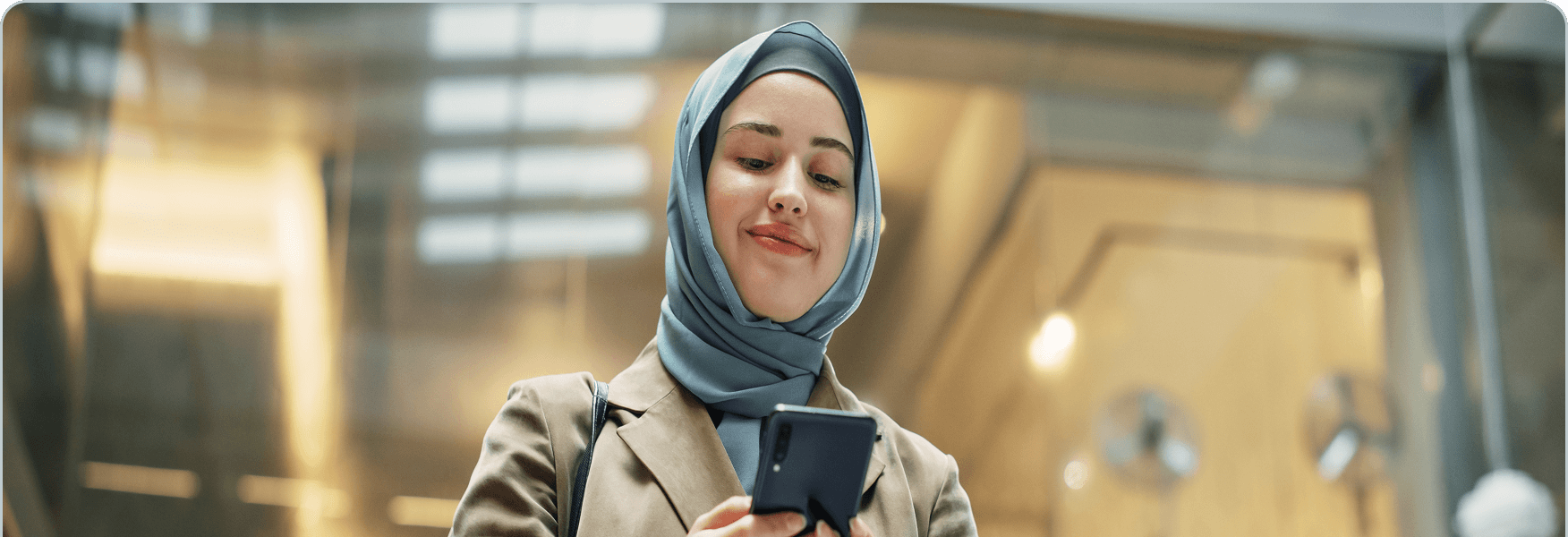 Una giovane donna con un hijab blu e una giacca beige sorride mentre guarda lo smartphone in un ambiente interno.