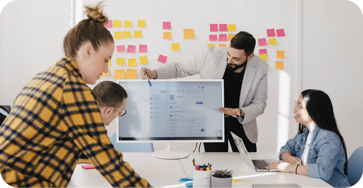 Vier Personen in einem Büro arbeiten gemeinsam um einen Computerbildschirm, mit bunten Haftnotizen an der Wand im Hintergrund.