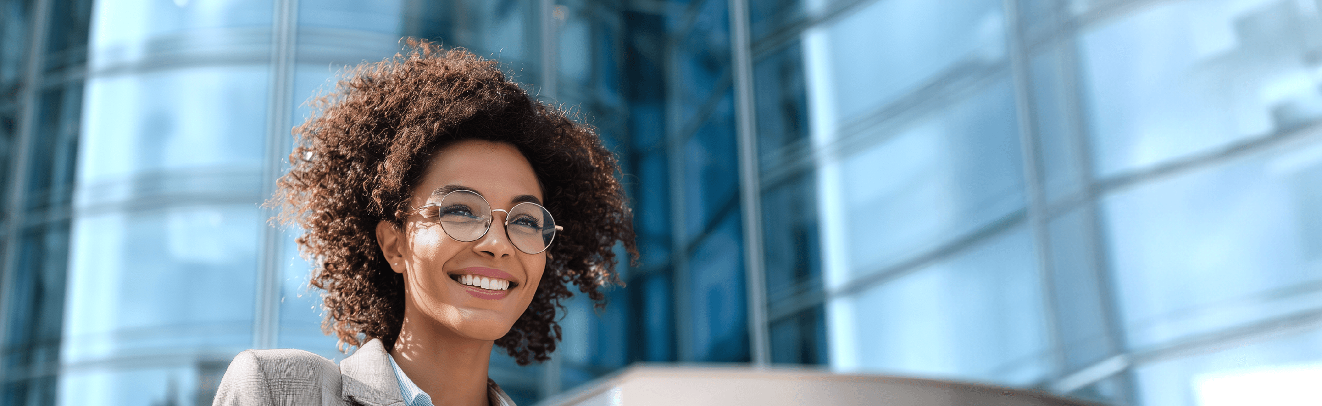 Profesional sonriente de pelo rizado y gafas que sostiene un dispositivo móvil; al fondo, un moderno edificio de oficinas