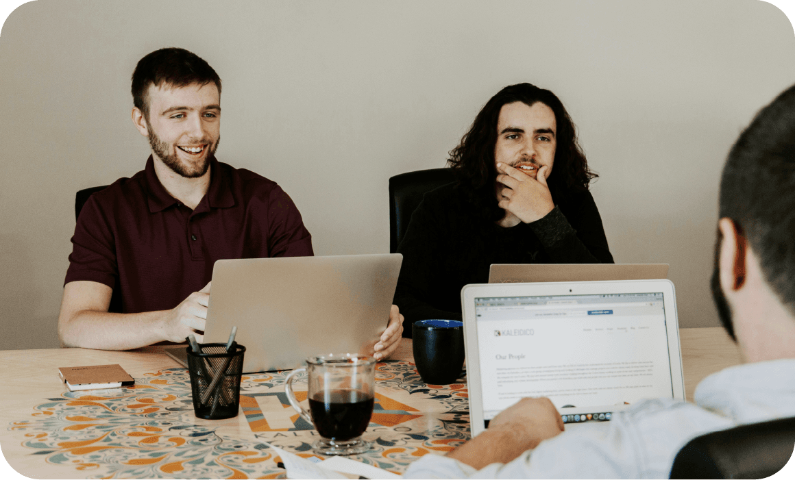 Drei Personen sitzen an einem Tisch mit Laptops und Gläsern und sind in eine Diskussion oder Besprechung vertieft.